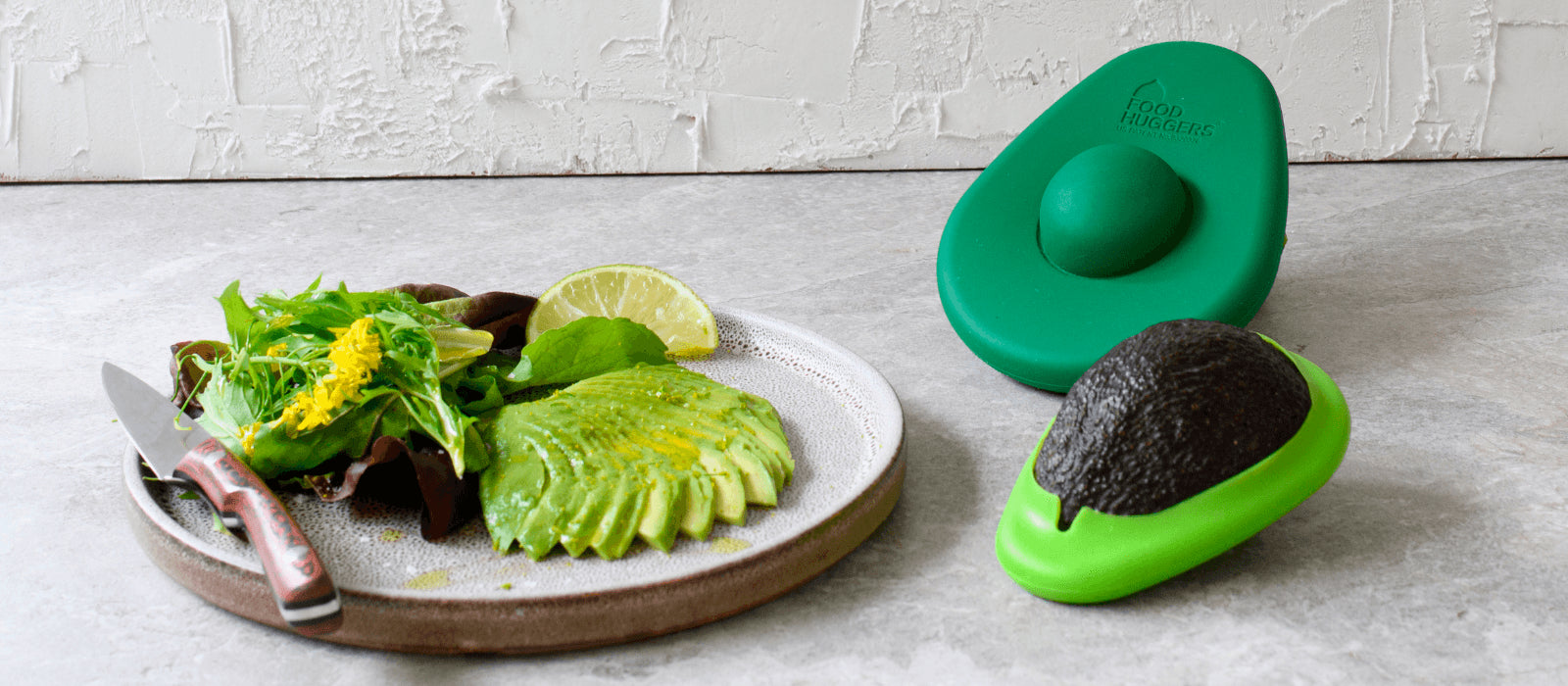 Two avocado huggers preserving an avocado, on a gray surface next to a plate with an avocado salad. Perfect for a sustainable kitchen.