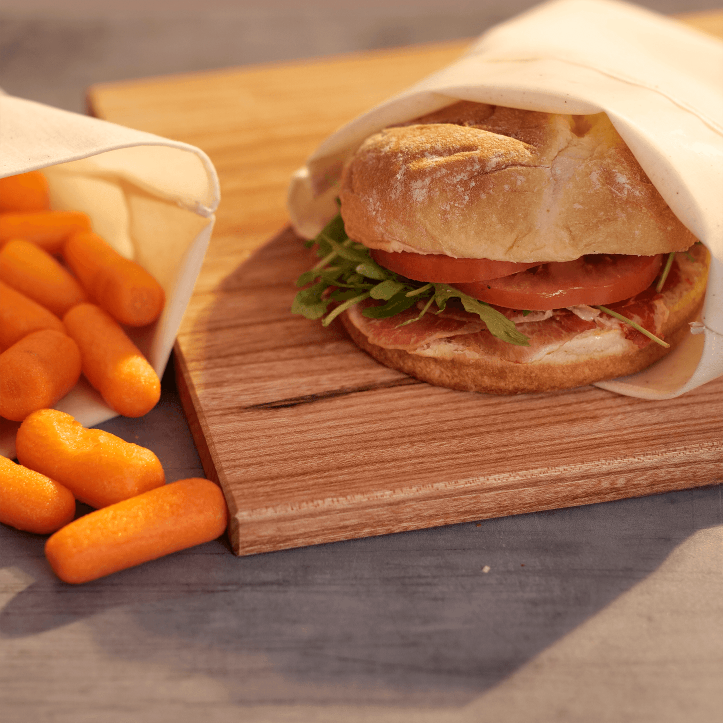 Food Huggers Fabric Bag preserving a sandwich on a wooden board next to another Food Hugger Fabric bag preserving carrot pieces