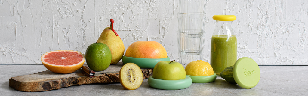 Five food huggers that preserve fruits for zero waste, on a gray surface next to a set of glasses.