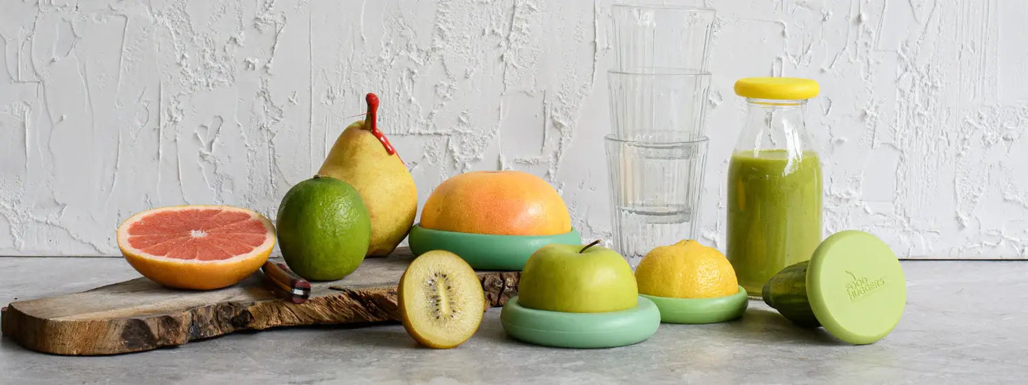 Five food huggers that preserve fruits for zero waste, on a gray surface next to a set of glasses and other fruits.