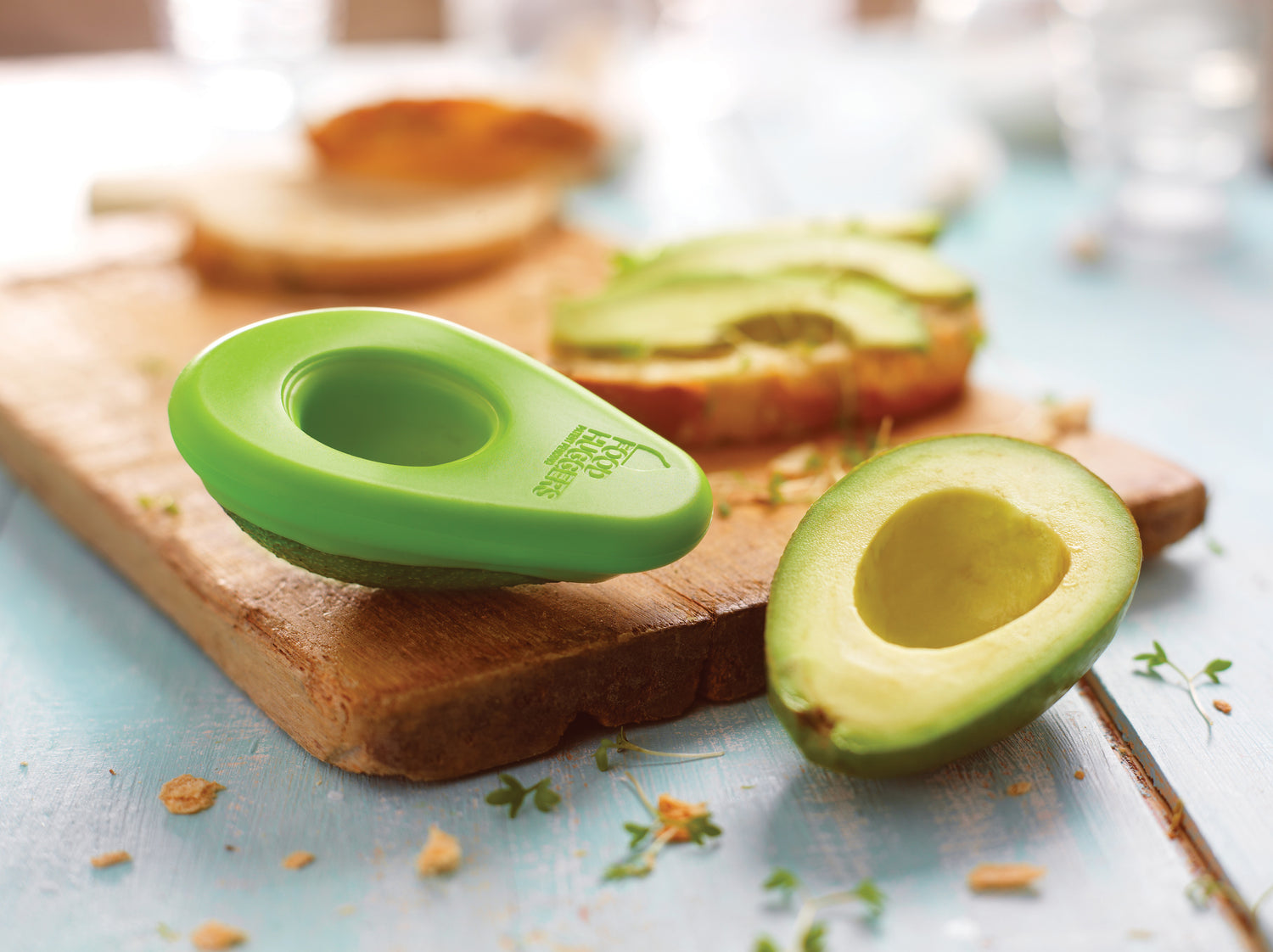 Half an avocado on a table, the other half on a wooden board protected by a green silicone wrap with the Food Huggers logo for zero waste.