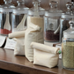 Three filled and closed Food Huggers Fabric Coffee Bag on a brown surface behind the bags glass vases filled with beans