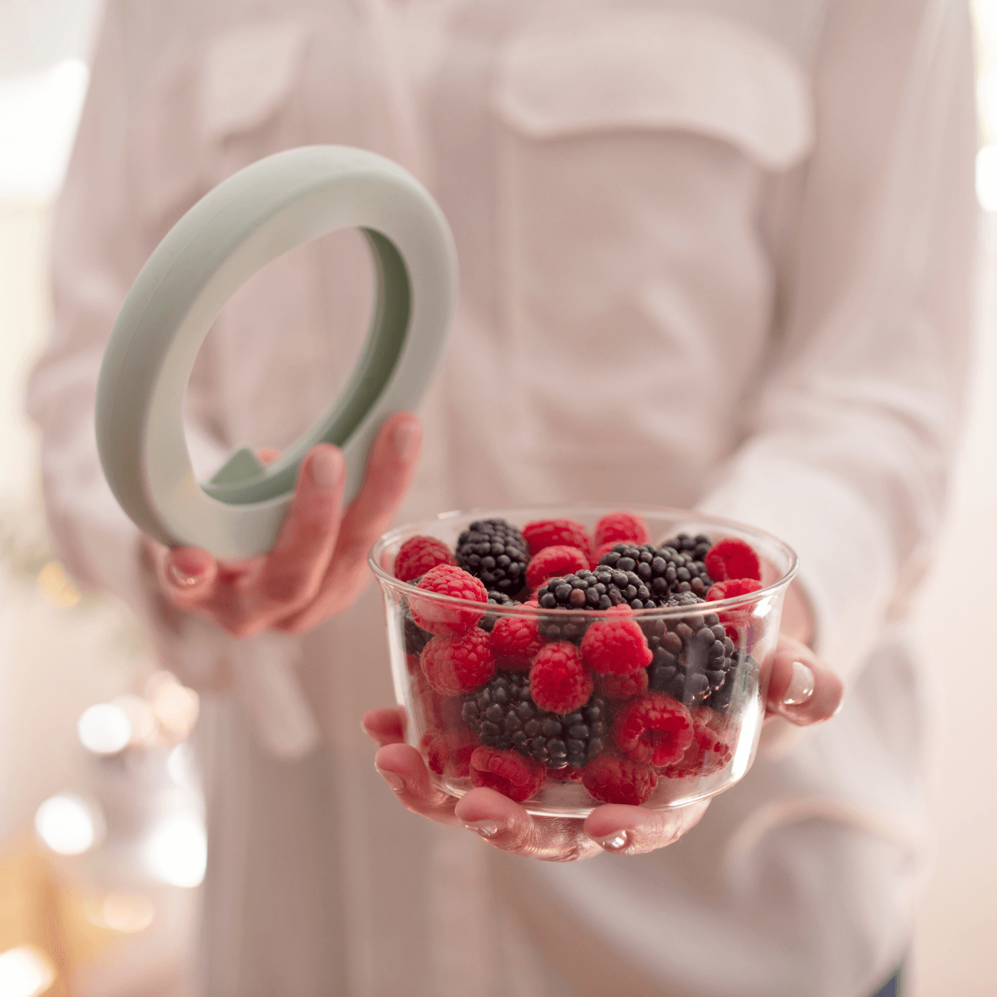 Woman holding in one hand a glass bowl filled with raspberries and in the other hand the adjustable food huggers lid